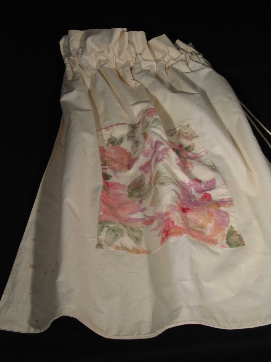 Hvit pose i lerret med løpegang og snor øverst. Den er dekorert på midten med et felt blomstret kretong.
(27 x 32 cm)