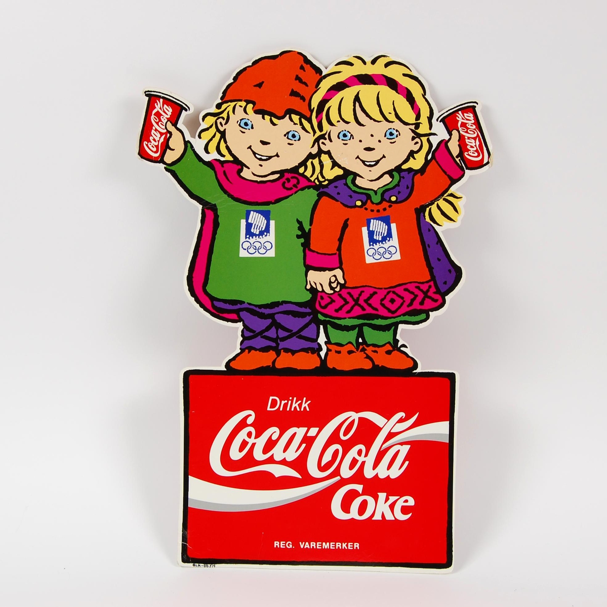 Reklameskilt med logo for Coca-Cola. Maskotene for de olympiske leker på Lillehammer i 1994, Kristin og Håkon, utgjør store deler av motivet og formen på skiltet. Skiltet er tosidig og likt på begge sider. Skiltet har noe slitasje.