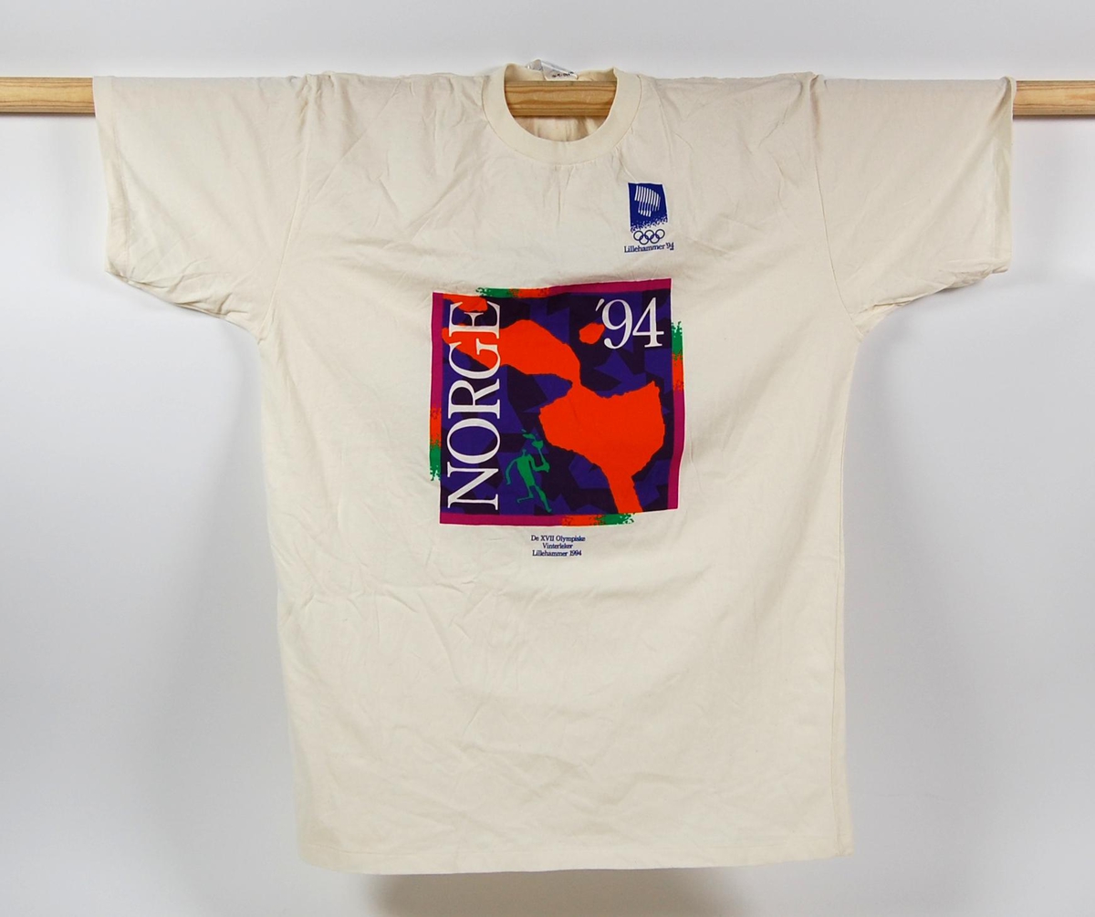 Gul/hvit t-skjorte i størrelse XL med flerfarget motiv av en fakkel og et piktogram av fakkelmannen. På t-skjorten er det også en logo for de olympiske vinterleker på Lillehammer i 1994.