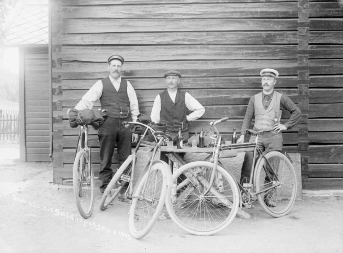 Kort: 04.07.1909. Sel, Otta. Gartner M. Skurdal med følge på tur til Galdhøpiggen. Gruppebilde, menn med sykler rundt et bord med drikkevarer.