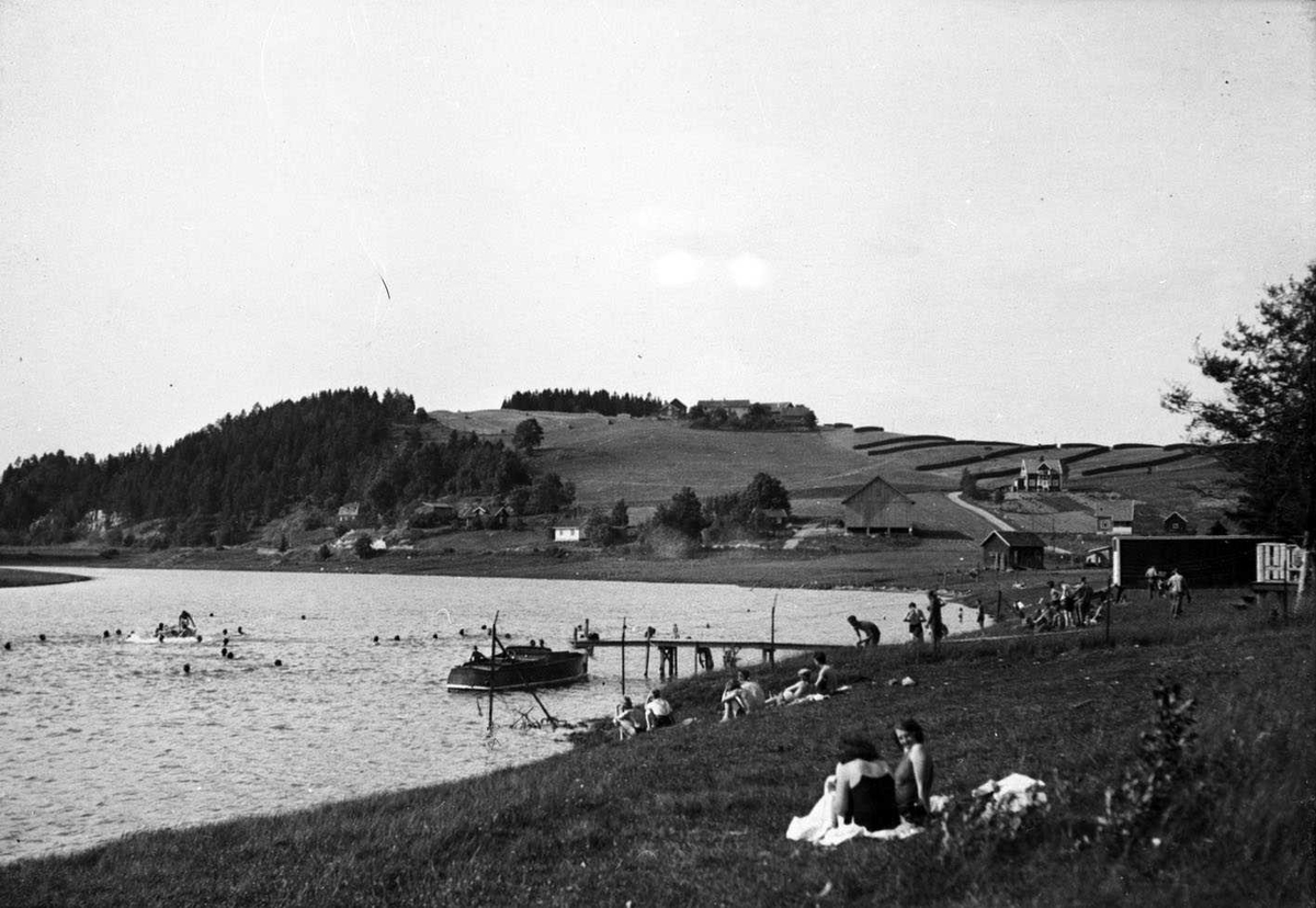 Soldatenes badeplass ved Nitelva ca. 1935
Nitteberg gård i bakgrunnen. Ei snekke ligger fortøyd ved brygga. I forgrunnen to kvinner i badedrakt. Elva er tett av badende.