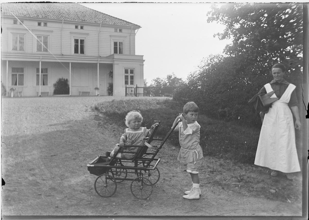 To barn leker med en vogn foran huset på Linderud Gård. En kvinne til høyre i bildet.