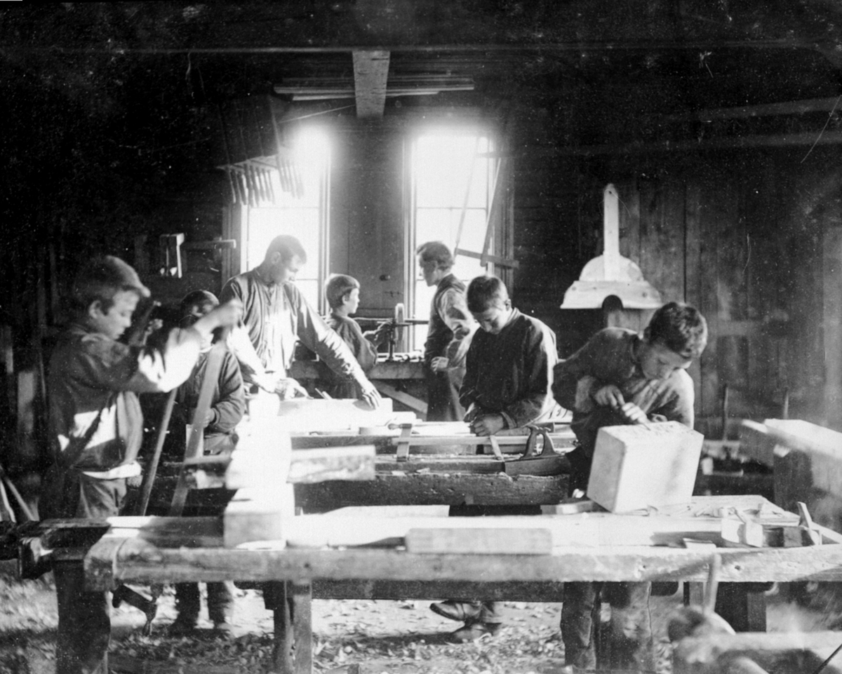 6 gutter arbeider i snekkerverkstedet på Toftes Gave, Nedre Sund, Helgøya. Lærer overvåker arbeidet.