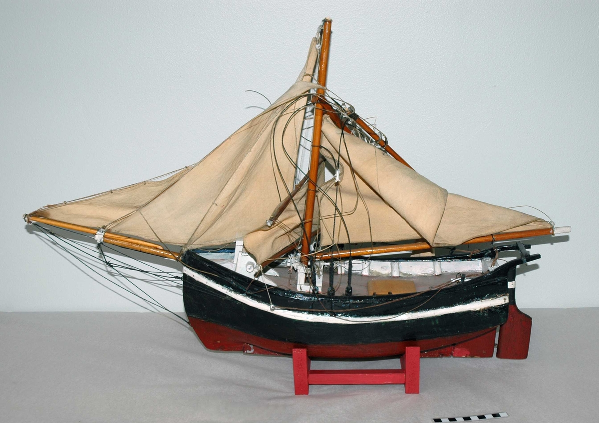 Båtmodell av en jakt, med ett mast og utslått seil. Masten er knekt rett av. Modellen hviler på et stativ av treverk.