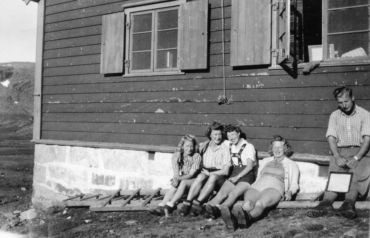 Fire kvinner og ein mann nyter sola i hytteveggen.