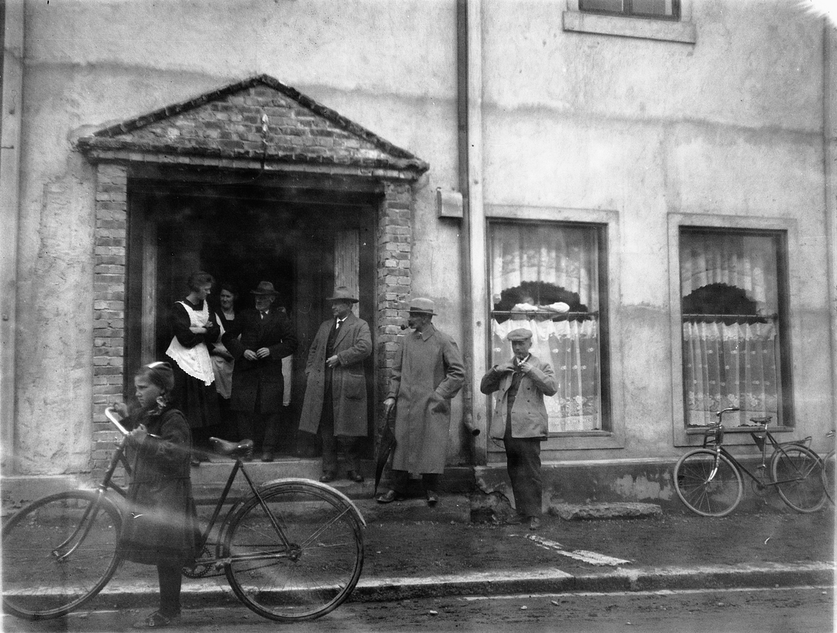 Gruppe mennesker ved inngangsdøra til en kafè. Jente med en sykkel i gata. 