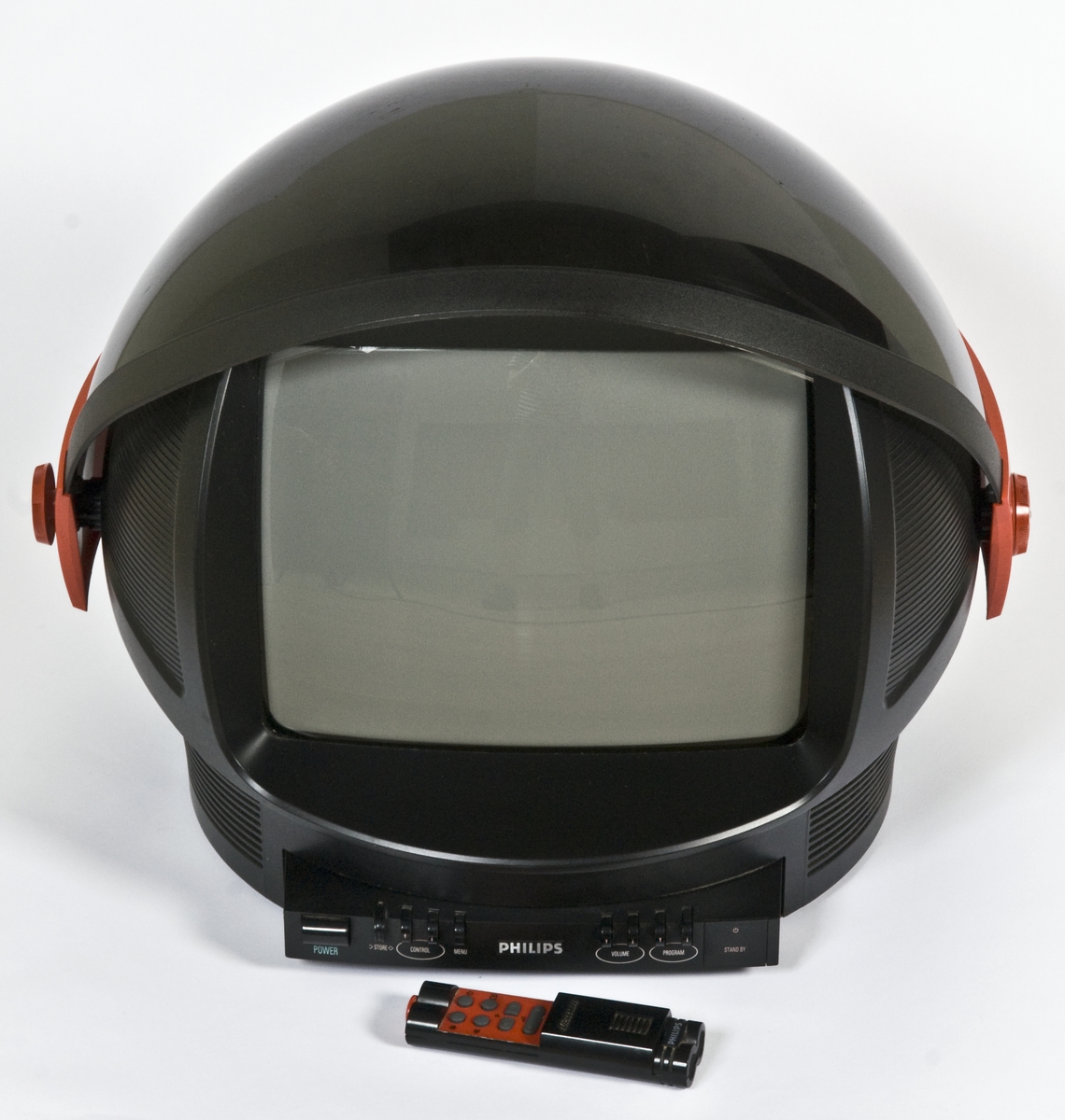Tv-apparat utformet som en astronaut-hjelm med hjul bak og vippbart visir i gjennomsiktig mørk plast. Fjernkontroll med skyvedeksel følger med.