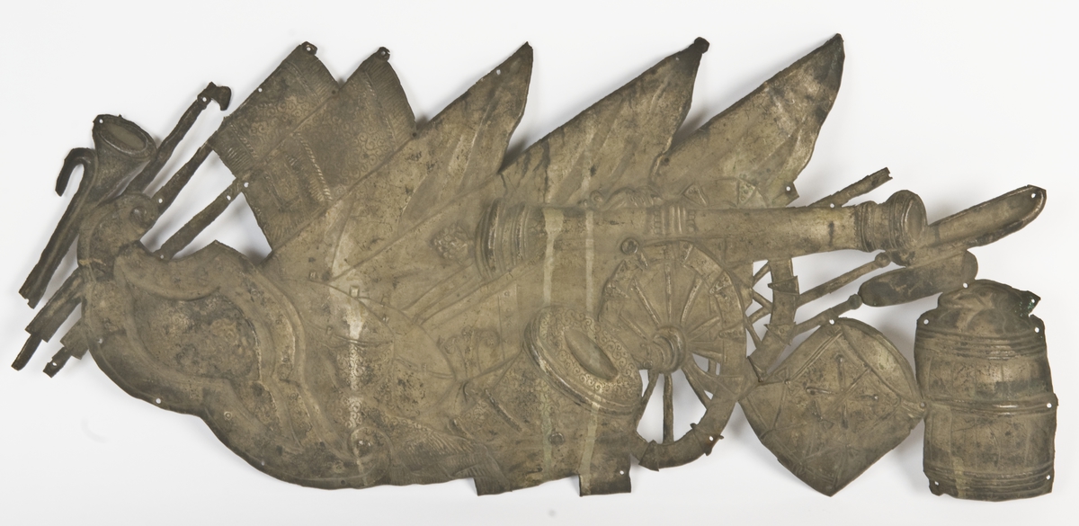 Beslag av hamret messingblikk, iflg. katalogkort fra likkiste. Barokkstil med våpenmotiver (økser, kanoner, trommer, faner etc.). Én av tre beslag med samme innføringsnummer fra Nordiska museet (NM.0063777AC).