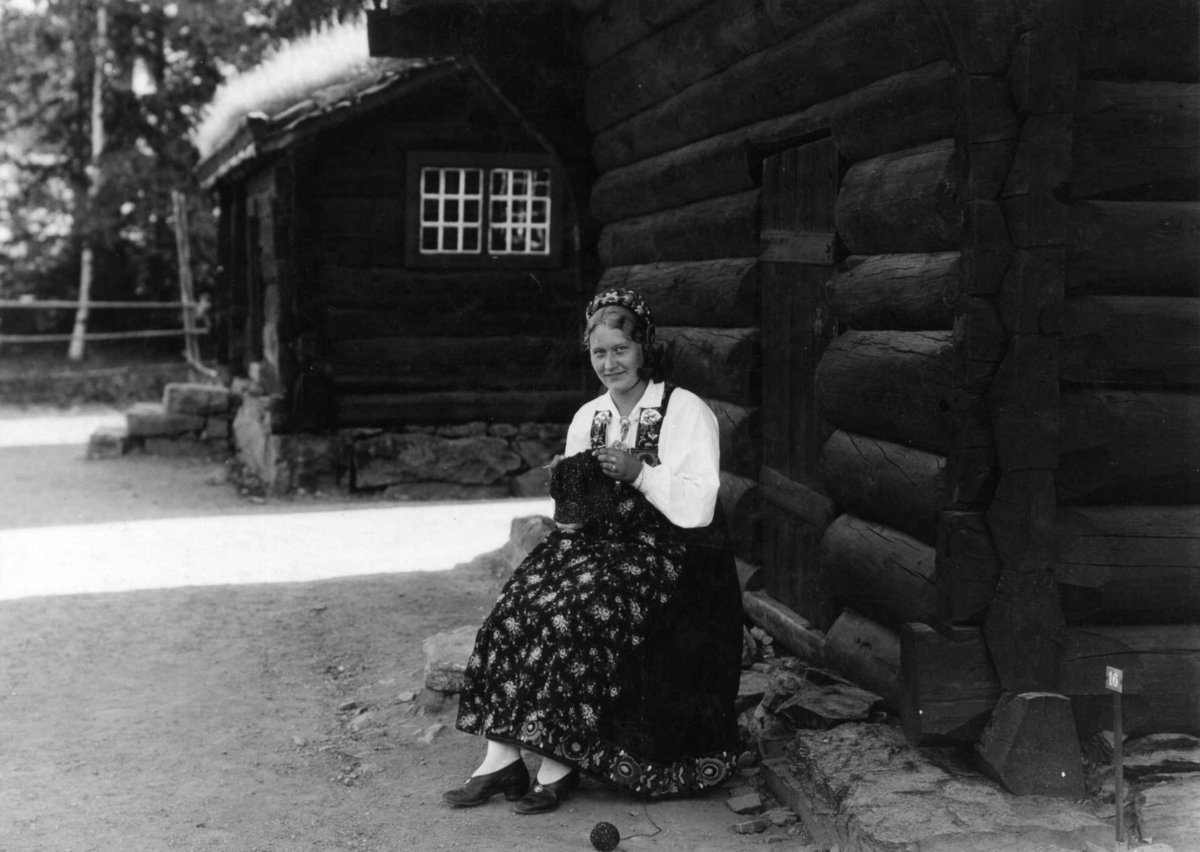 Vert, Hilda Gjerdene, i Hallingdalsdrakt i Hallingdalstunet på Norsk Folkemuseum. Hun sitter og strikker ved veggen til Loftstua, bygning nr. 112, med Bjørnebergstølstua, bygning nr. 113 i bakgrunnen.