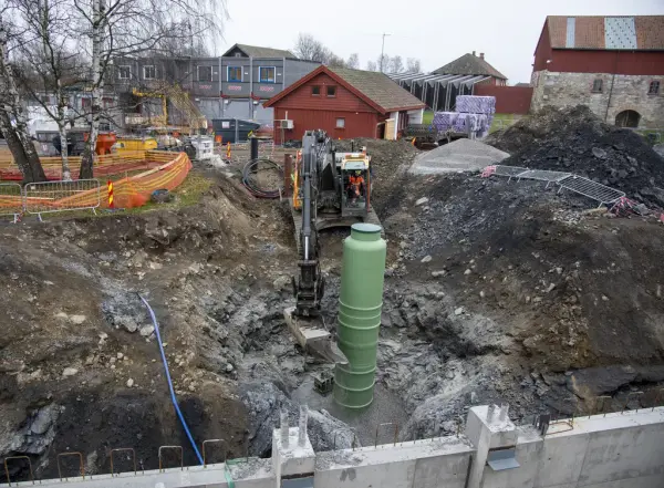 Gravemaskin står nede i byggegrop og graver på plass et tykt, grønt rør.