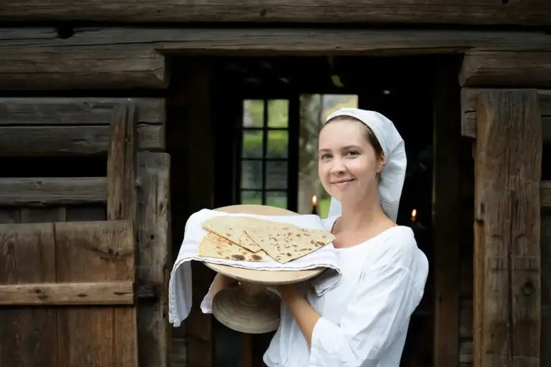 Kvinne i historisk drakt holder fat med nystekte lefser foran tømmerbygning i Numedal på Norsk Folkemuseum.